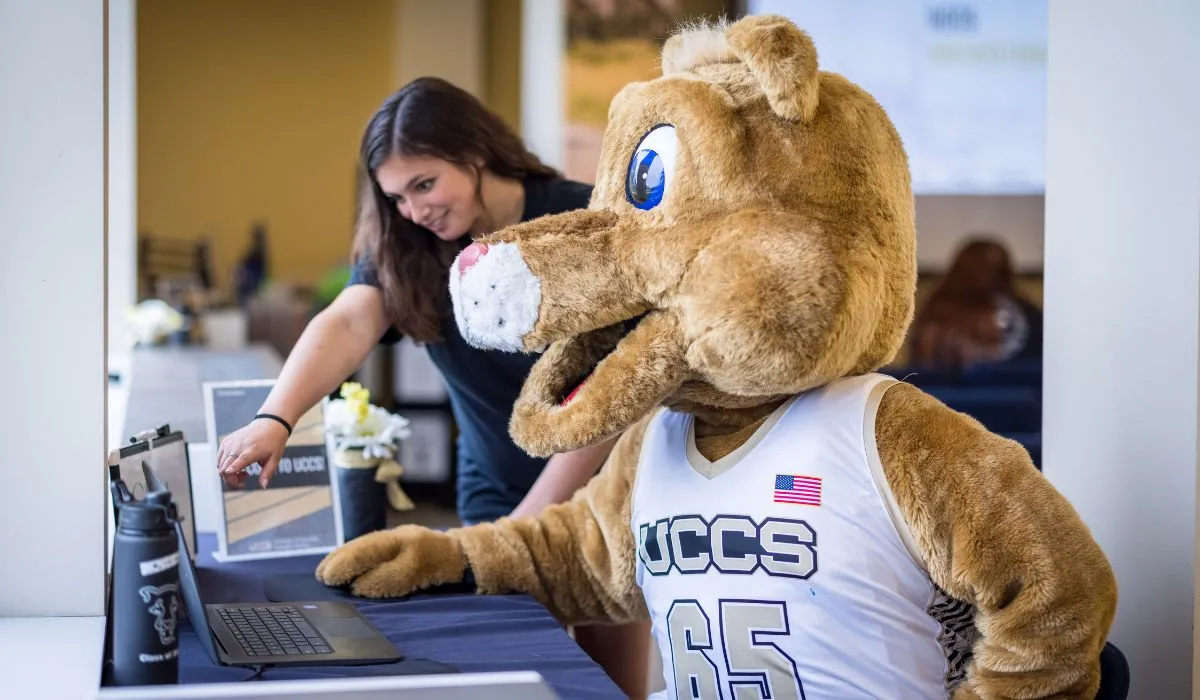 UCCS Mascot, Clyde, sitting with an Admissions Counselor on the computer